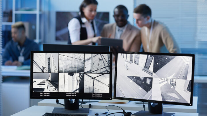 Office desk with two monitors displaying black-and-white security camera footage; keyboard, mouse, and papers on the desk.